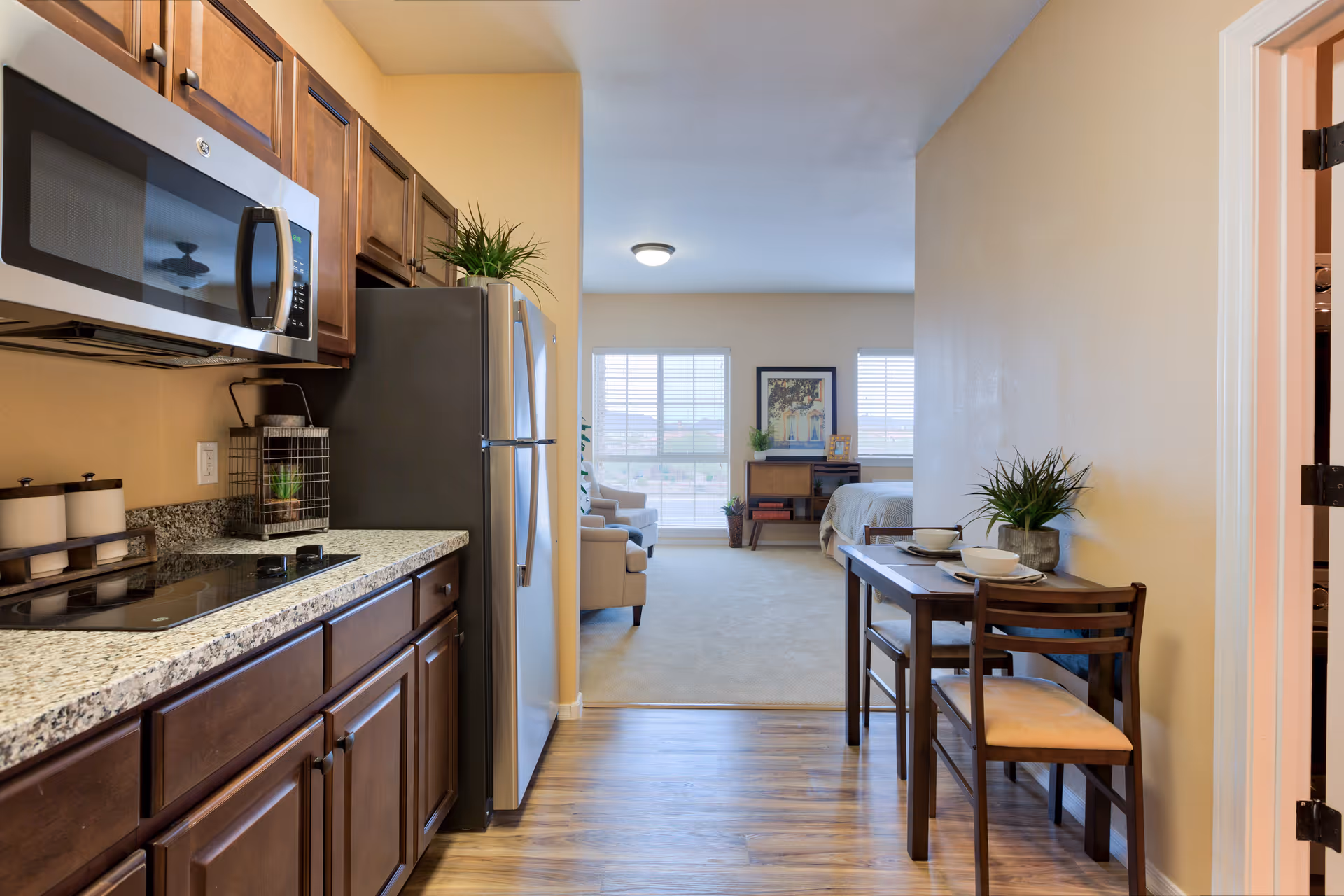 View of a kitchen area with wooden cabinets, granite countertops, a microwave, and a refrigerator on the left. On the right, there is a small dining table set for two with a plant centerpiece. Beyond the kitchen and dining area, a living space with a beige armchair, a bed, and large windows letting in natural light is visible.