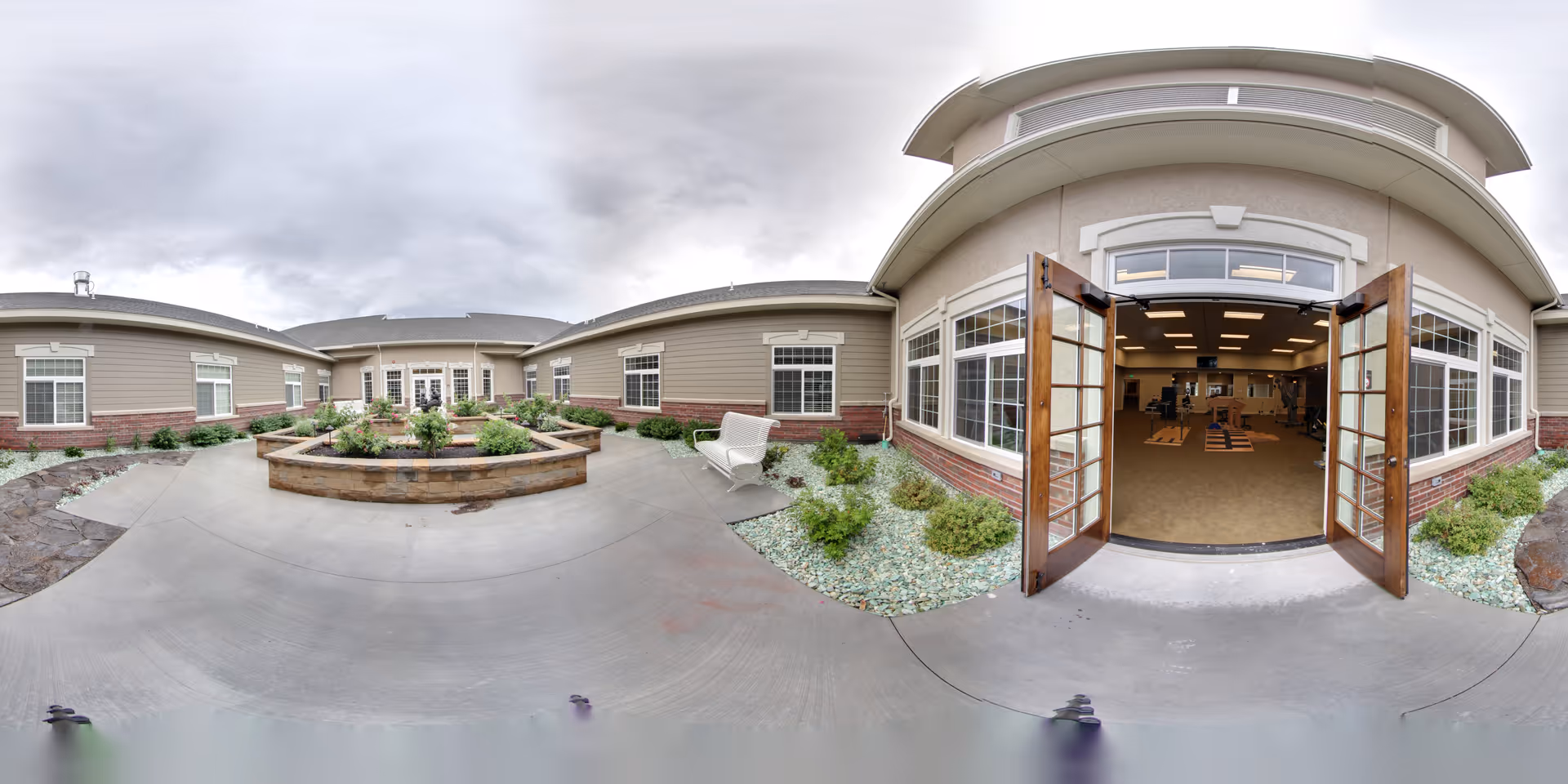 Outdoor courtyard area of Carriage Cove Short Stay Rehabilitation facility featuring a central raised garden bed with plants, surrounded by a concrete walkway. The building has beige siding with white trim and multiple windows. On the right side, double wooden doors are open, revealing an interior room with exercise equipment. There is a white bench on the right side near the garden bed, and landscaping with small bushes and rocks along the building walls. The sky is overcast.