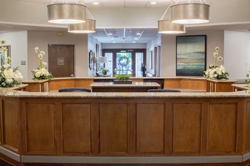 Wooden reception desk in a nursing facility lobby with pendant lights, floral arrangements, and a view toward the front entrance.