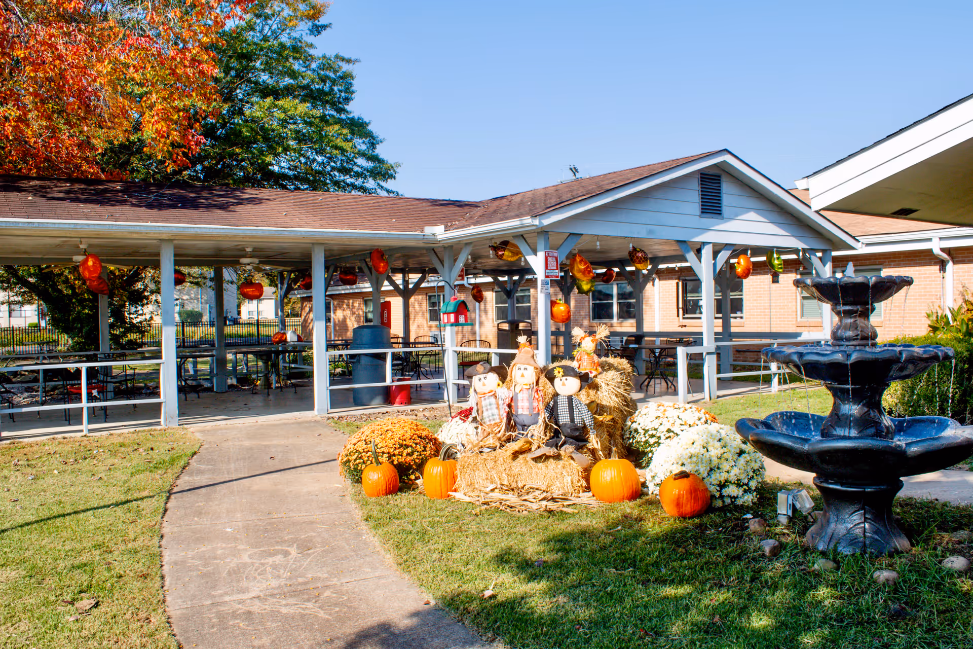 Outdoor covered patio and courtyard with fall decorations—pumpkins, hay bales, scarecrows—and a tiered fountain in front of a brick rehabilitation center.