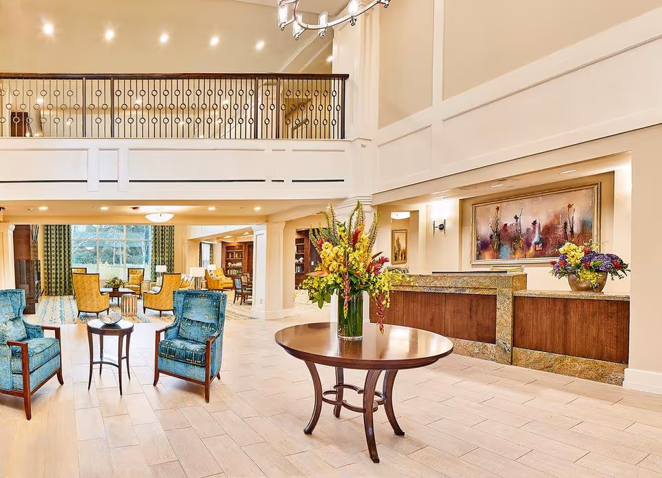 A spacious and well-lit senior living facility lobby with a round wooden table in the center holding a large vase of colorful flowers. To the left, there are two blue upholstered armchairs with a small round table between them. In the background, there are more seating areas with yellow chairs and a bookshelf. The reception desk is on the right side with another flower arrangement and a large framed artwork on the wall behind it. The area features high ceilings, recessed lighting, and a second-floor balcony with decorative railing.