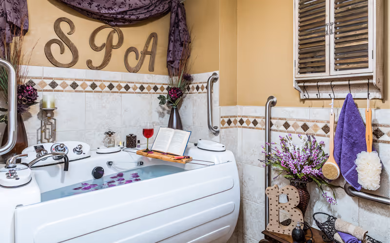 Decorative spa-style bathroom featuring a white soaking tub with a tray holding a book and wine glass, 'SPA' wall letters, purple accents, flowers, and grab bars.