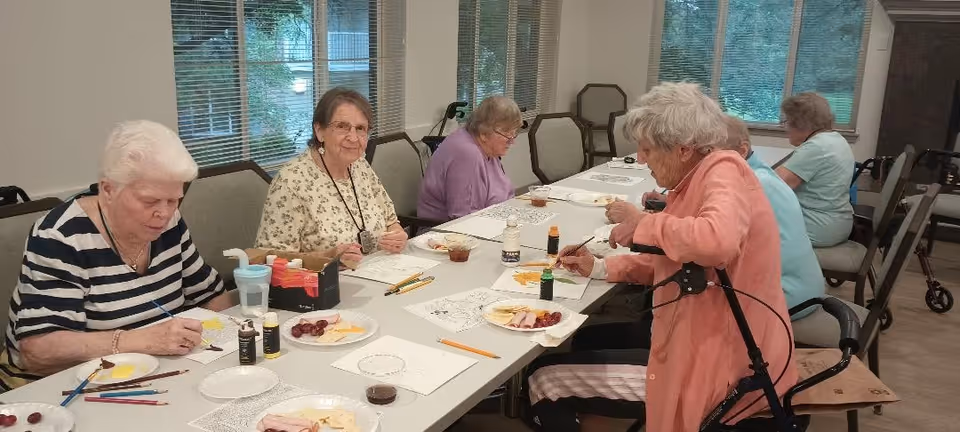 A group of elderly women seated around a long table engaged in painting and coloring activities. The table has art supplies, colored pencils, paint bottles, and plates with snacks. The room has large windows with blinds and a view of greenery outside.