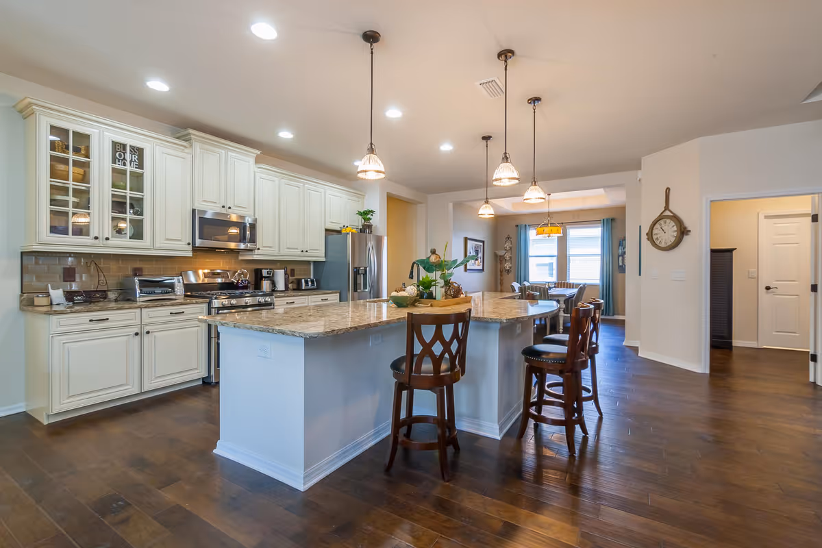 Bright open kitchen with a large marble island, pendant lights, bar stools, white cabinets, stainless appliances, and hardwood floors.