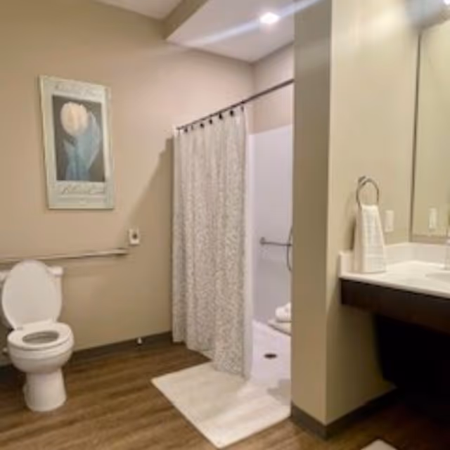 A clean bathroom with a toilet, a shower with a patterned curtain, a white bath mat, a sink with a towel hanging on a ring, and a framed floral artwork on the wall. The floor is wood-style and the walls are beige.