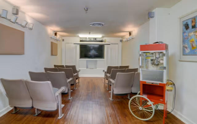 A small theater or media room with rows of gray chairs facing a large flat-screen TV mounted on a white wall. The room has wooden flooring and white walls with light fixtures. On the right side, there is a red and silver popcorn machine with a large wheel. A bulletin board with photos is visible on the right wall.