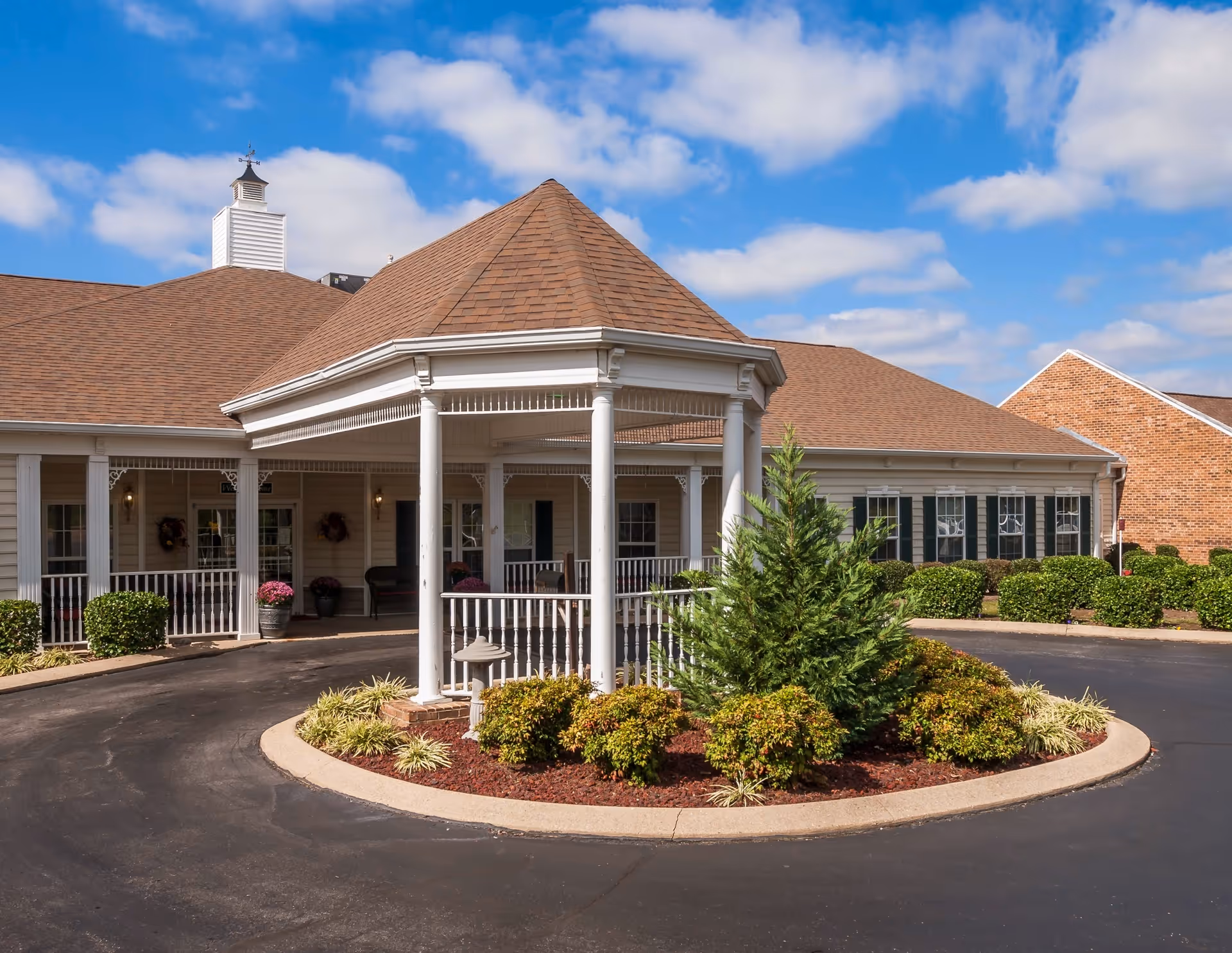 Front exterior view of Charter Senior Living of Paris building with a covered entrance, circular driveway, landscaped bushes, and a clear blue sky with some clouds.