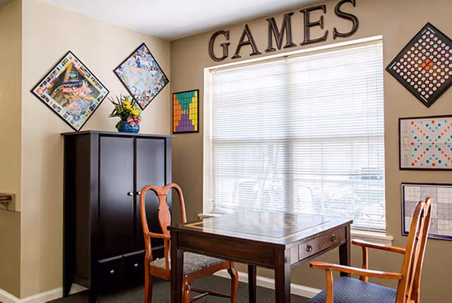 Small games room with a wooden table and chairs, a storage cabinet, wall-mounted game boards, and a large window beneath a 'GAMES' sign.