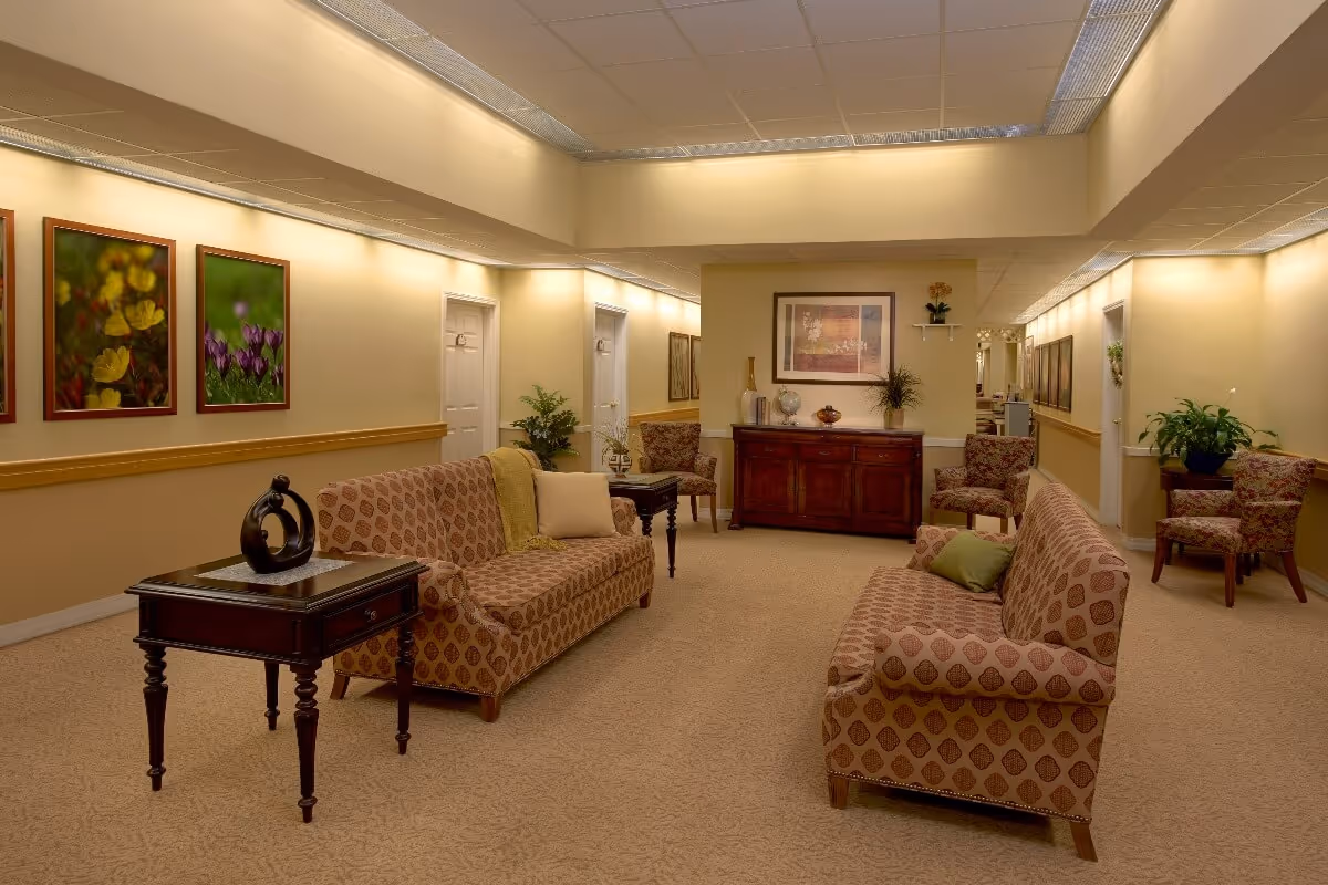 A well-lit sitting area in a senior living facility with two patterned sofas facing each other, a dark wooden side table with a decorative sculpture, framed floral artwork on the walls, several potted plants, and a wooden cabinet with decorative items and a framed picture above it.