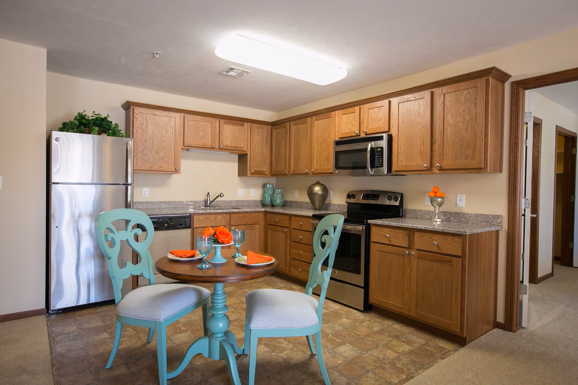 A kitchen area in a retirement facility featuring wooden cabinets, a stainless steel refrigerator, stove, and microwave. There is a small round wooden table with two turquoise chairs, set with orange napkins, blue glasses, and a centerpiece of orange flowers. The floor is tiled, and there is a doorway leading to another room.