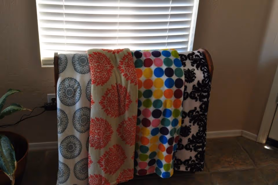 Four colorful patterned blankets hanging over a wooden rack in a room with a window covered by blinds in the background. A potted plant is partially visible on the left side, and the floor is tiled.