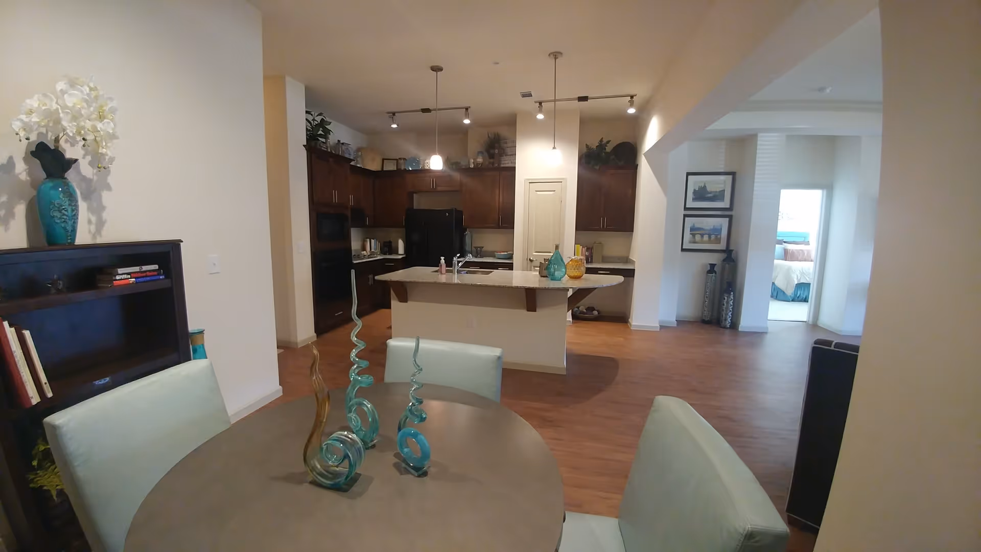 Interior view of a senior living facility showing a dining area with a round table and four chairs, decorative glass sculptures on the table, a kitchen with dark wood cabinets and a granite countertop island, and a glimpse into a bedroom with a bed and framed pictures on the wall.