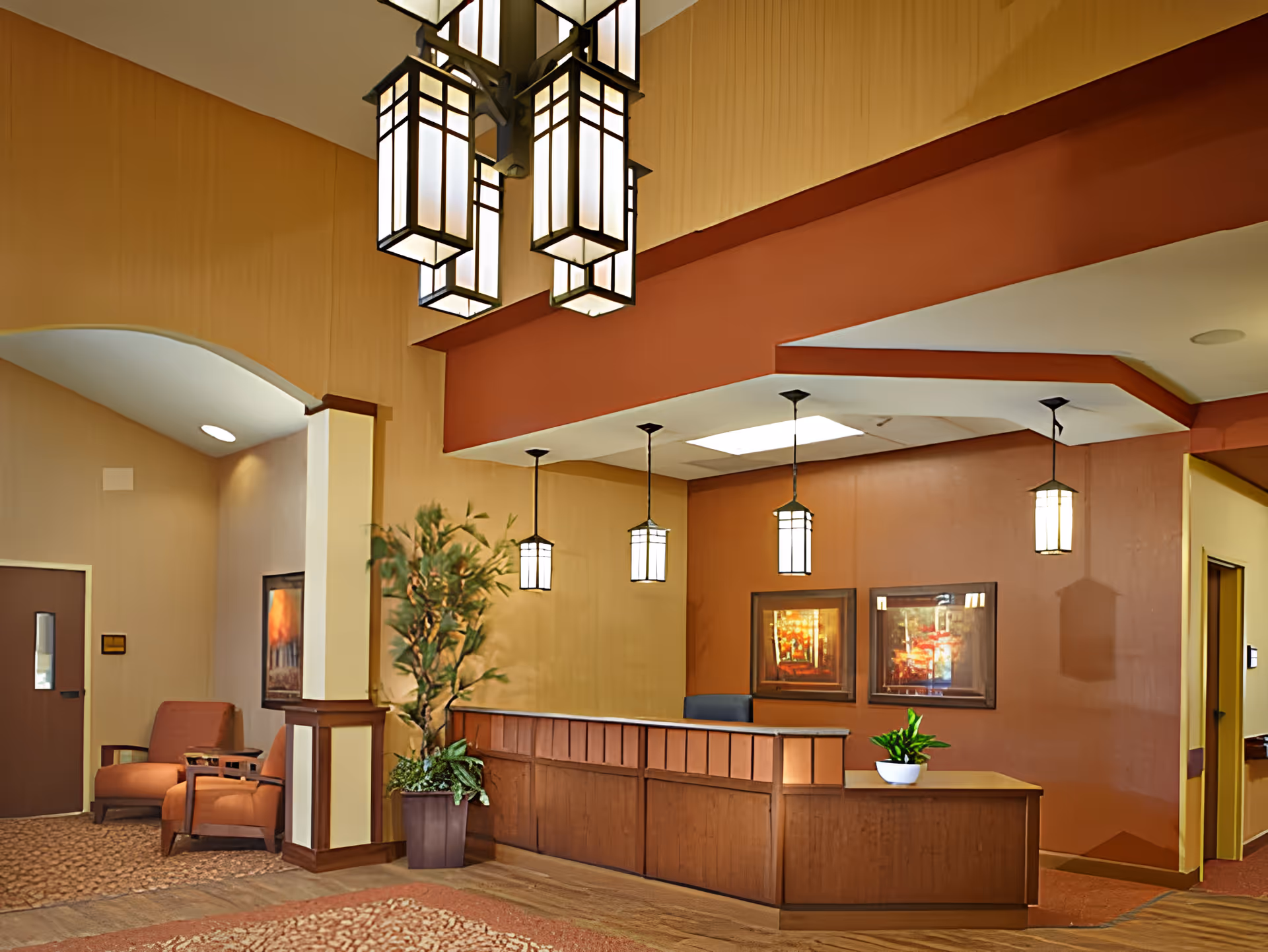 Lobby reception area with a wooden front desk, hanging pendant lights, seating, and potted plants.
