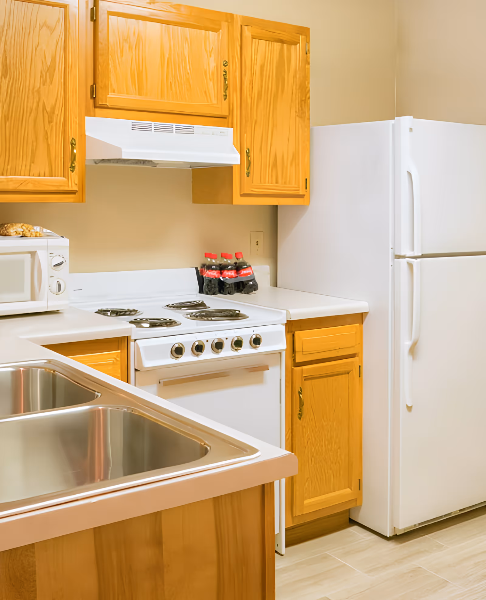 A kitchen area with wooden cabinets, a white electric stove with four burners, a white refrigerator, a microwave, and a double stainless steel sink. There are three bottles of soda placed on the countertop next to the stove.