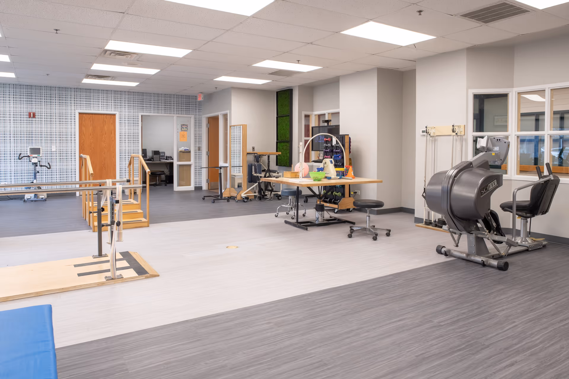 A spacious rehabilitation or physical therapy room with exercise equipment including parallel bars, a seated exercise machine, therapy tables with various therapy tools, and a small set of stairs with handrails. The room has a mix of light and dark wood flooring, white walls, and a ceiling with fluorescent lights. There are doors and windows leading to adjacent rooms.