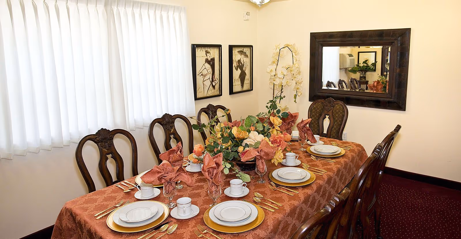 A formal dining room with a long table covered in a patterned rust-colored tablecloth, set with white plates on gold chargers, white cups and saucers, gold cutlery, and rust-colored folded napkins in glasses. The table has a centerpiece of artificial flowers in shades of yellow, orange, and green. The room has wooden chairs with ornate backs, a large mirror on one wall, two framed vintage-style fashion prints on another wall, and white curtains covering the window.
