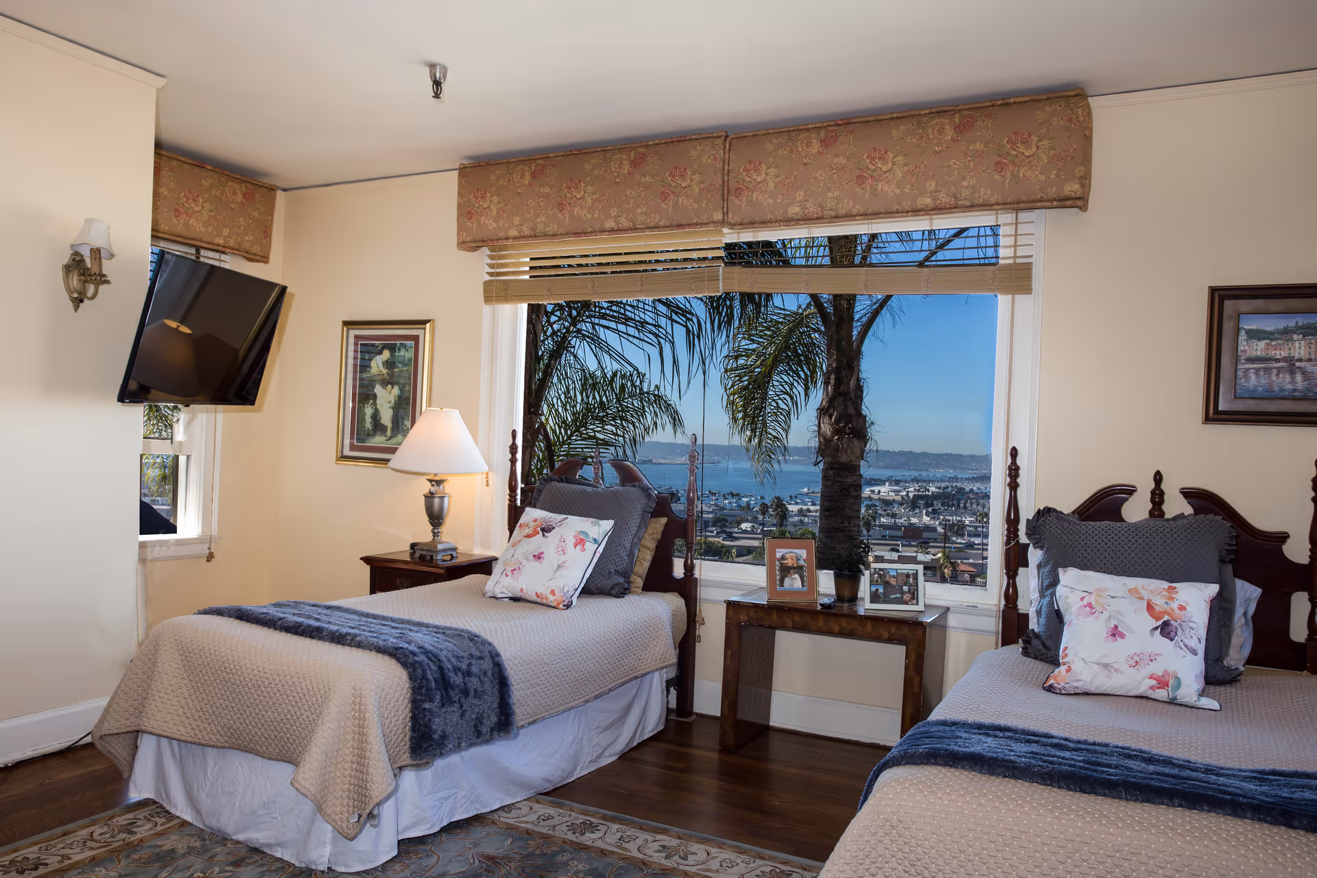 A bright bedroom with two twin beds, each with beige bedspreads, floral pillows, and blue throws. Between the beds is a wooden table with framed photos and a potted plant. A large window behind the table offers a view of palm trees and a cityscape with water in the distance. A wall-mounted TV is on the left wall, and a lamp sits on a nightstand between the left bed and the window. The room has floral valances above the windows and framed artwork on the walls.