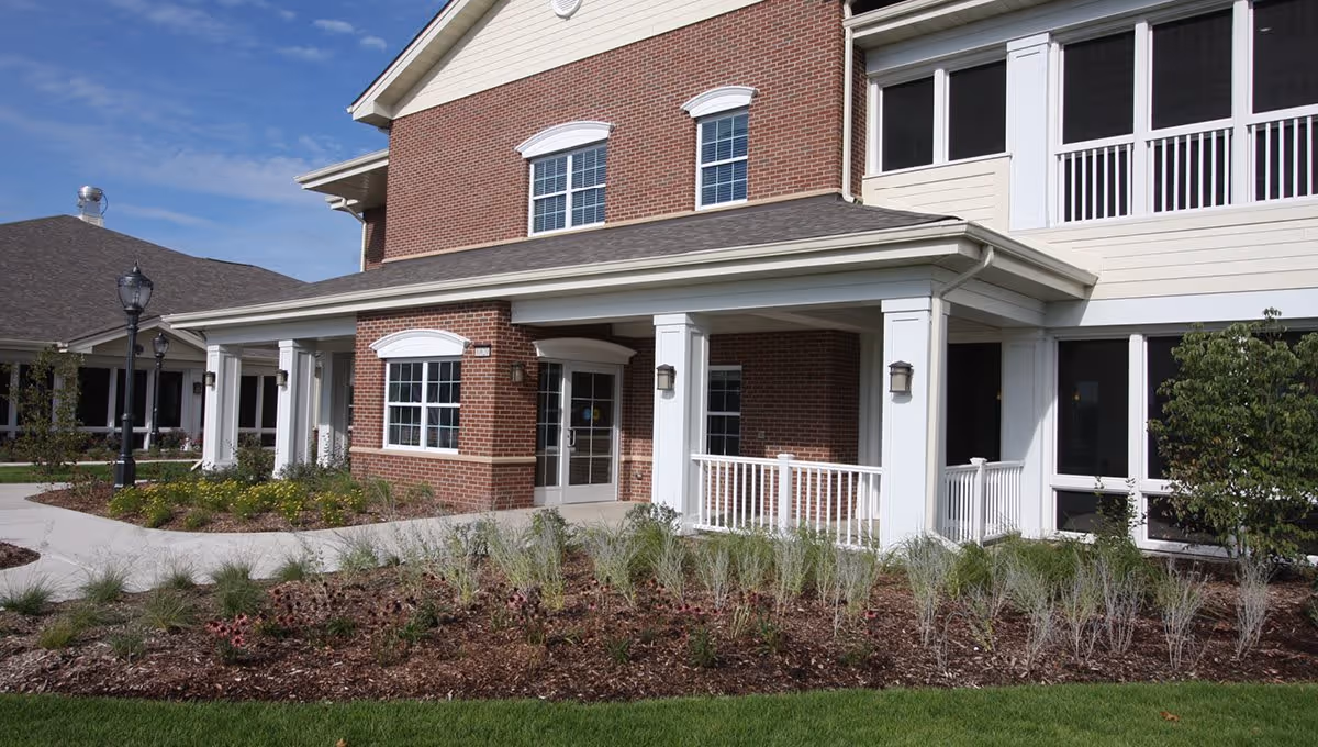 Exterior view of a brick and white siding building with a covered entrance supported by white columns, surrounded by landscaped garden beds and green grass under a clear blue sky.