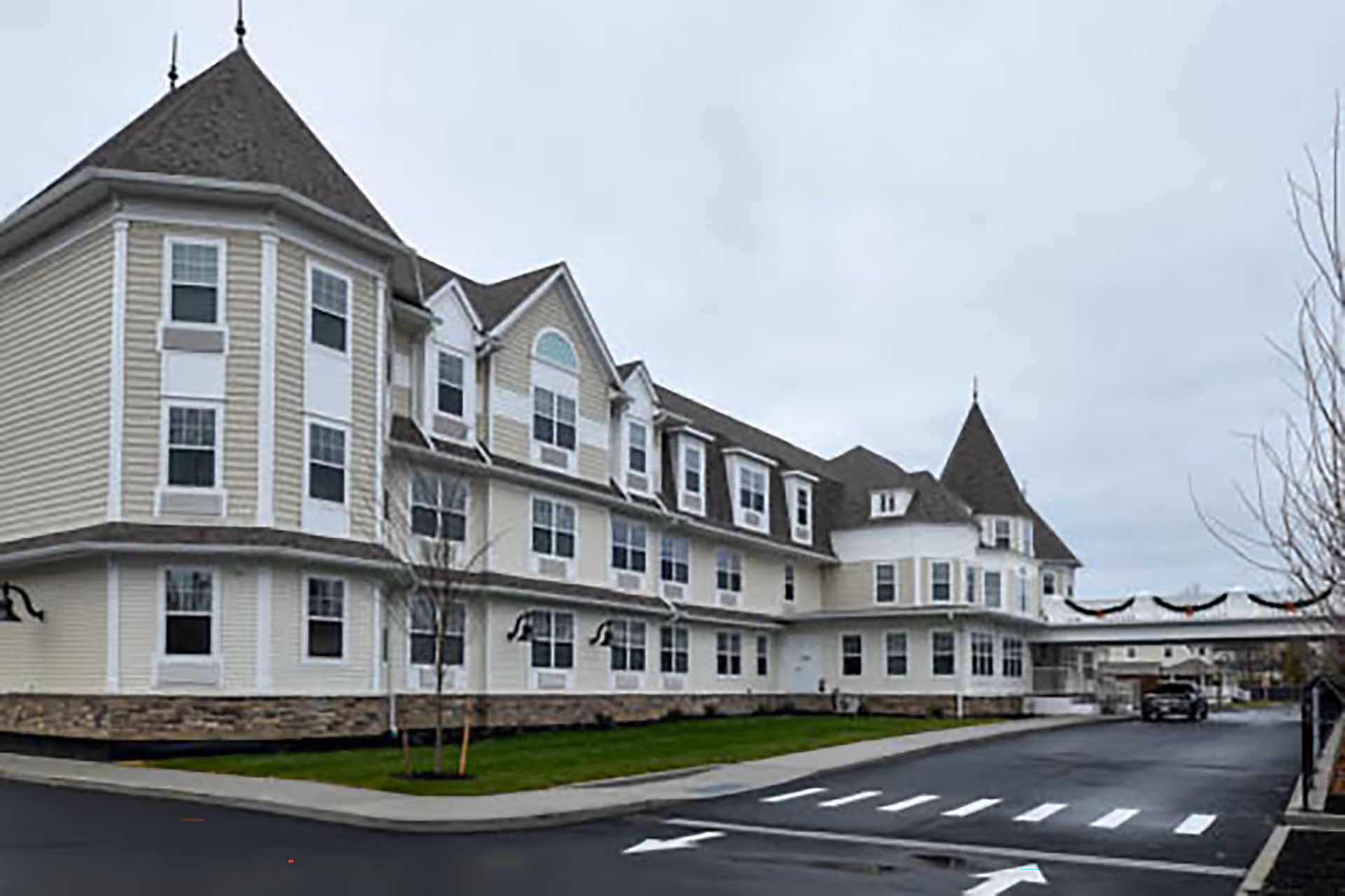 Exterior view of a large, multi-story senior living facility building with beige siding, white trim, and multiple windows. The building features a peaked roof and a covered driveway entrance. There is a paved road with crosswalk markings and some small trees and grass areas in front of the building under a cloudy sky.