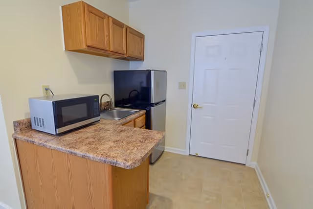Small kitchen area with a countertop, microwave, stainless steel mini refrigerator, and a small sink. There are wooden cabinets above and below the countertop. A white door with a gold handle is visible on the right side of the image.