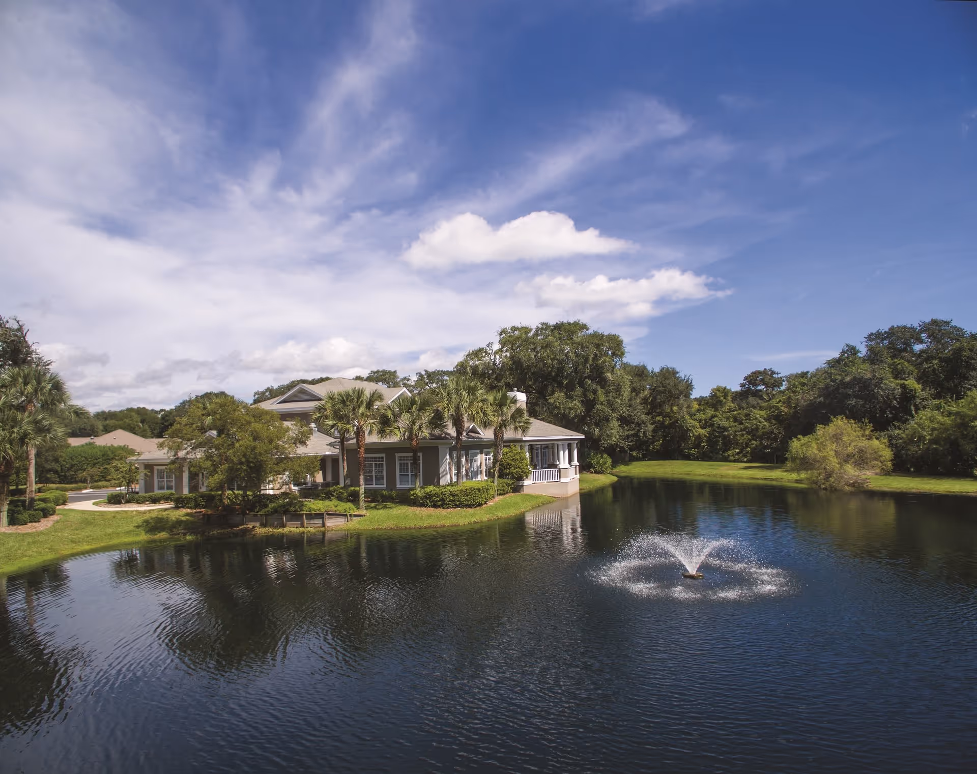 A serene outdoor scene at Retreat At Osprey Village featuring a large pond with a water fountain in the center, surrounded by lush greenery and trees. A building with a porch and palm trees is visible on the edge of the pond under a partly cloudy blue sky.