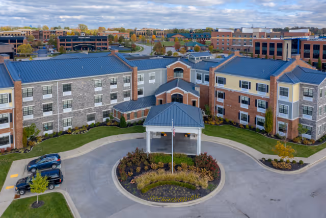 Aerial view of a senior living facility with a circular driveway and landscaped center island featuring shrubs and flowers. The building has multiple stories with a combination of brick and stone exterior walls, and a blue metal roof. There is an entrance canopy with an American flag in front. Surrounding the facility are parking areas, green lawns, and other buildings in the background under a partly cloudy sky.