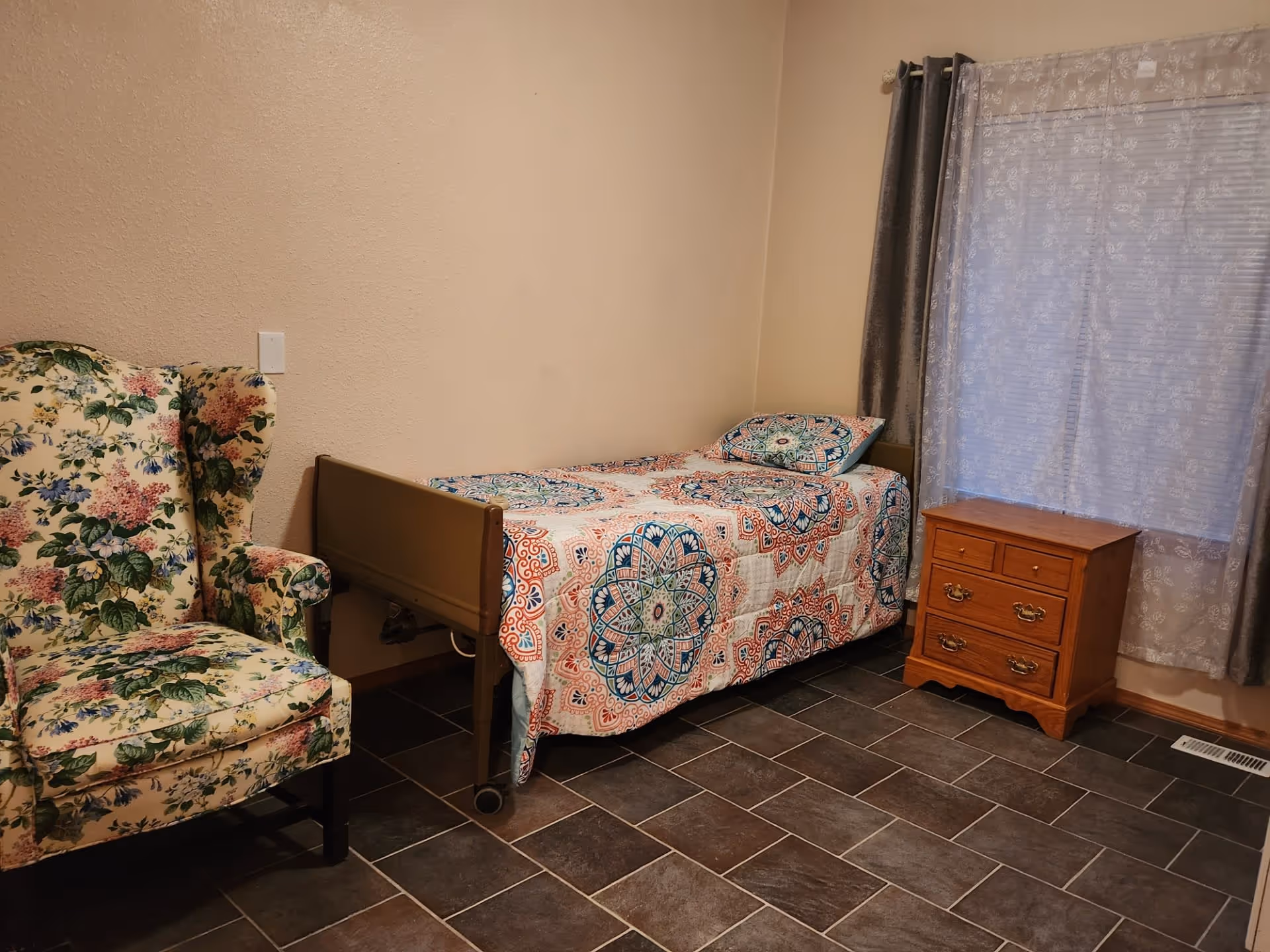 A small bedroom with a single bed covered in a colorful patterned quilt and matching pillow. Next to the bed is a wooden nightstand with drawers. To the left of the bed is a floral upholstered armchair. The room has tiled flooring and a window with sheer white curtains and gray drapes.
