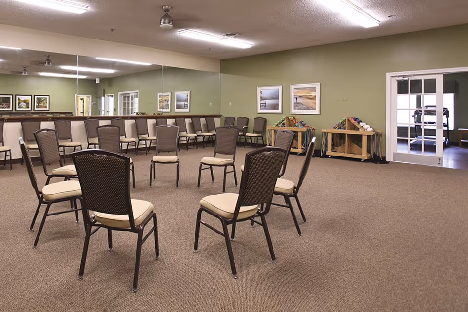 A spacious activity room with chairs arranged in a circle, a mirrored wall, framed artwork, and exercise equipment along a green wall.