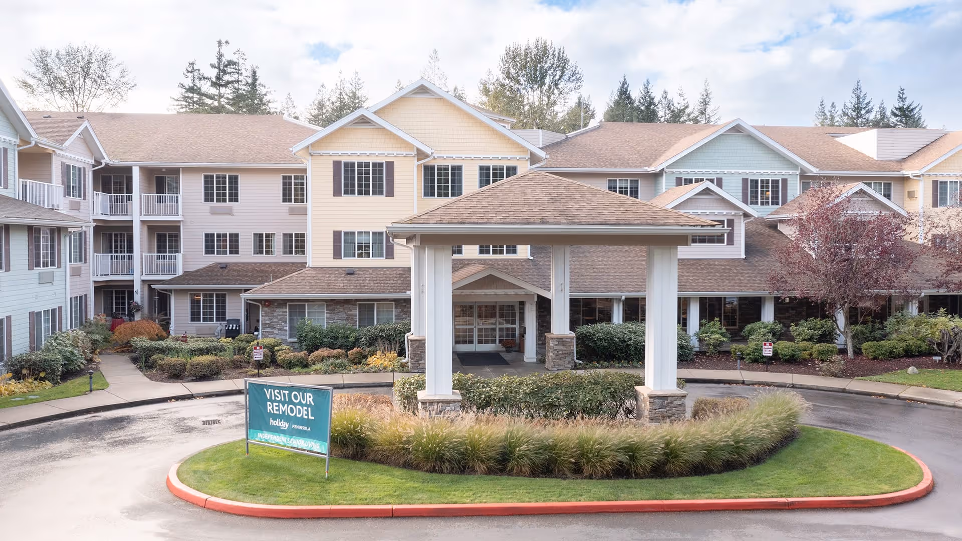 Front exterior view of the Holiday Peninsula senior living facility showing a multi-story building with beige, yellow, and light blue siding, a covered entrance with white pillars, landscaped greenery, and a sign that reads 'Visit Our Remodel Holiday Peninsula'.