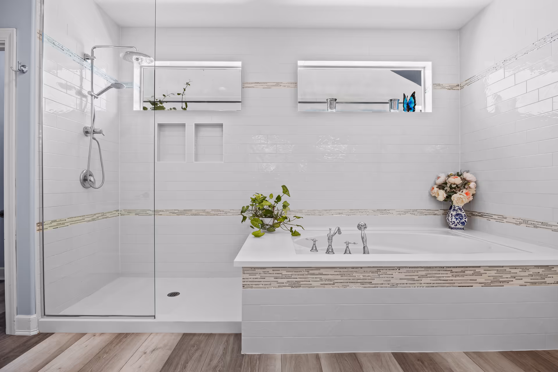 Bright modern bathroom with a glass-enclosed shower, a large soaking tub, decorative tile trim, and plants and flowers on the tub ledge.