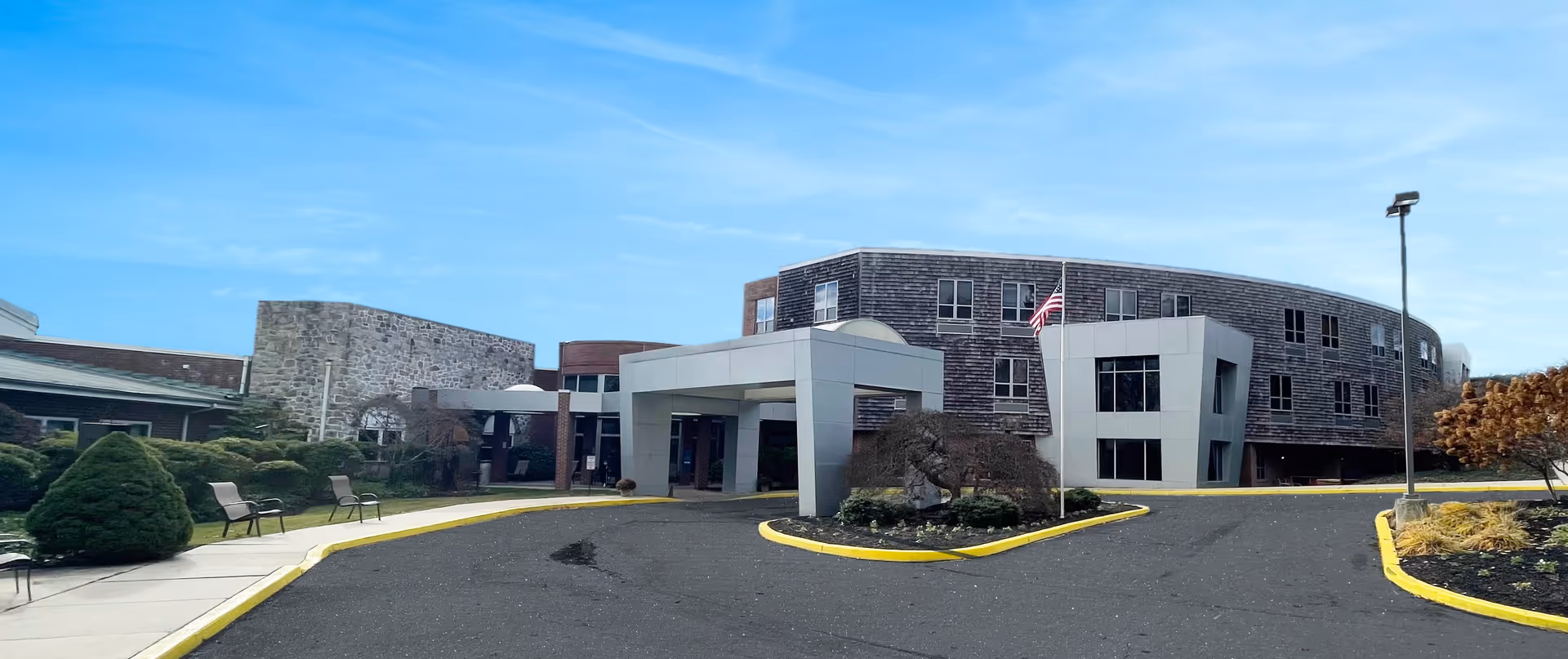 Exterior view of Springfield Crossings senior living facility showing a curved multi-story building with a covered entrance, an American flag, landscaped bushes, benches along the sidewalk, and a clear blue sky.