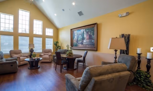 A cozy living room with several cushioned armchairs and a round wooden table with four chairs. The room has large windows with blinds, a tall plant, a table lamp, and a large framed painting on a yellow wall. The floor is wooden.