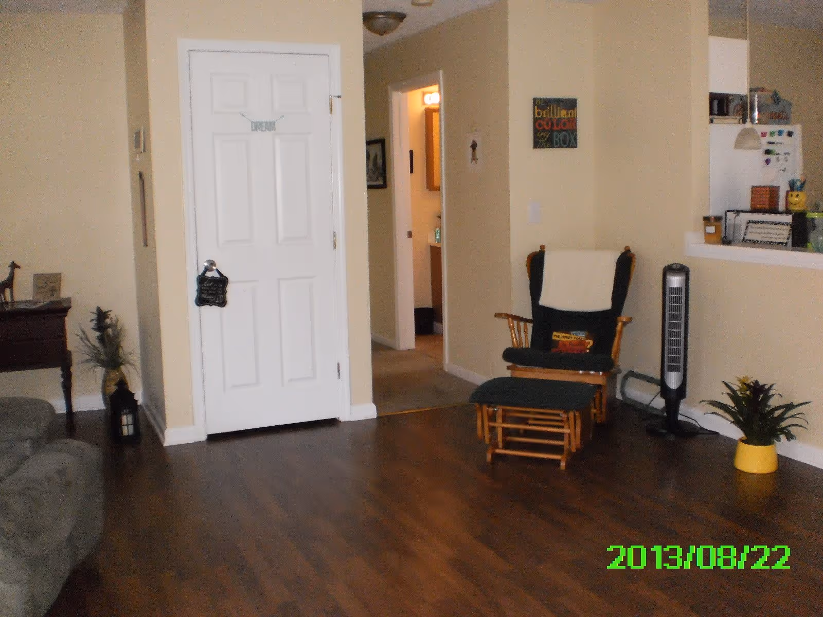 Interior living room with wood floors, a cushioned glider chair and ottoman, a white door opening to a hallway, a tower fan and potted plants.