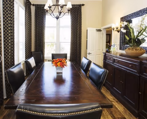 A dining room with a long wooden table surrounded by eight black leather chairs. A vase with orange and red flowers is centered on the table. The room has large windows with patterned dark curtains, a chandelier overhead, and a wooden sideboard with a large mirror and a decorative plant on top. The walls are light-colored and the floor is wooden.