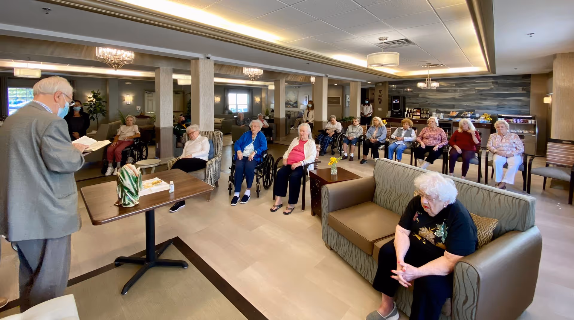 A group of elderly residents seated in a spacious, well-lit common area of an assisted living facility. An elderly man wearing a mask stands at a table reading from a book, while the residents listen attentively. The room features comfortable chairs, sofas, and a snack area in the background.