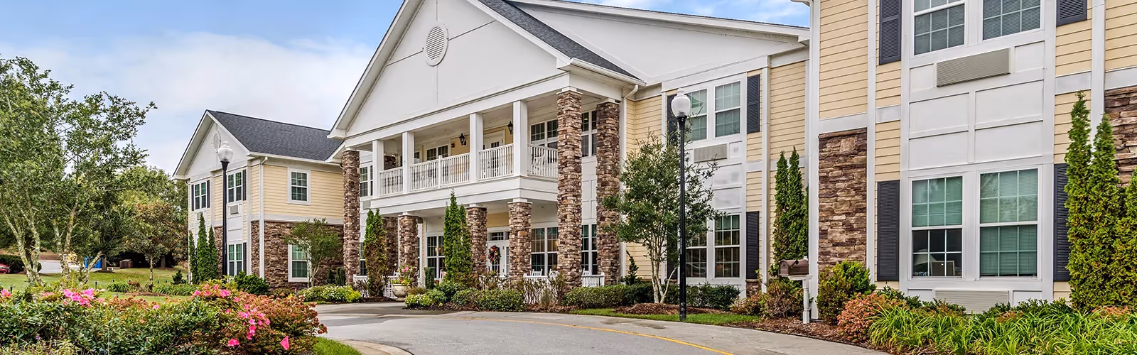 Exterior view of a senior living facility building with beige siding, stone pillars, white trim, and multiple windows. The building has a covered porch with white railings and columns. There are landscaped bushes, flowers, and trees along the driveway leading to the entrance under a partly cloudy sky.
