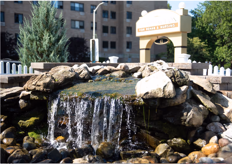 A small artificial waterfall flowing over rocks in a landscaped outdoor area with a building and a sign for rehabilitation and nursing in the background.