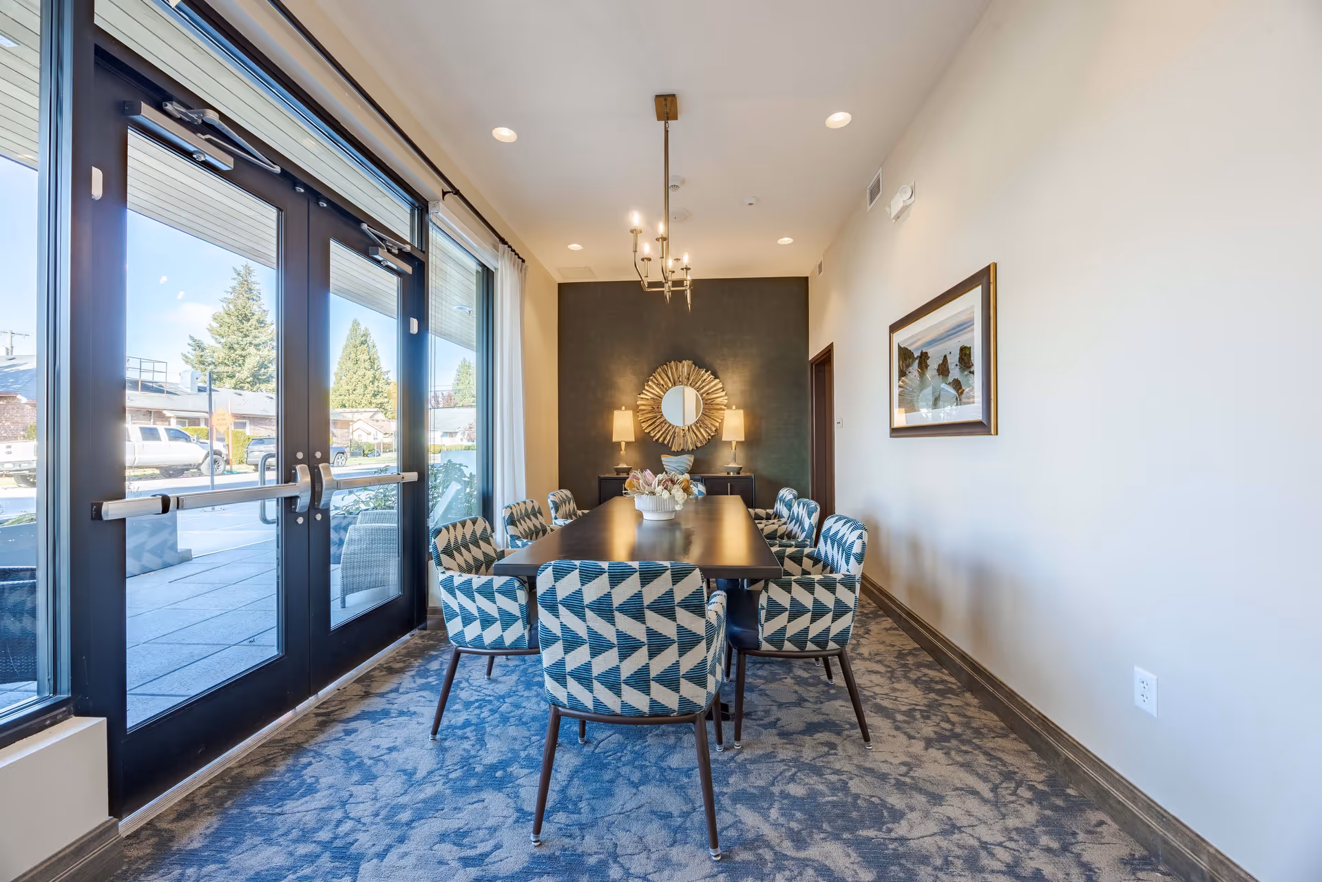 Bright dining/meeting room with a long table surrounded by patterned chairs and large glass doors to the outside.