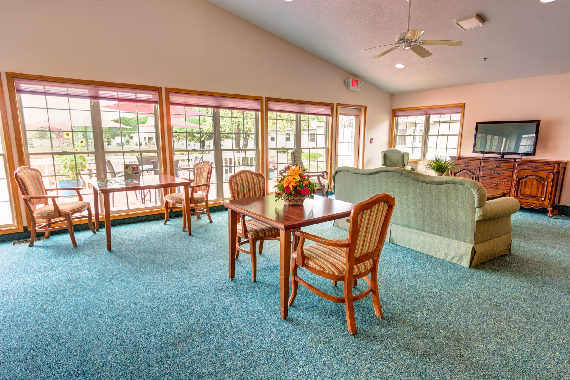 A bright and spacious common area in a senior living facility with large windows letting in natural light. The room features a green upholstered sofa and armchair, wooden tables with striped cushioned chairs, a wooden cabinet with a flat-screen TV on top, and a ceiling fan. Outside the windows, there is a patio area with umbrellas and greenery.