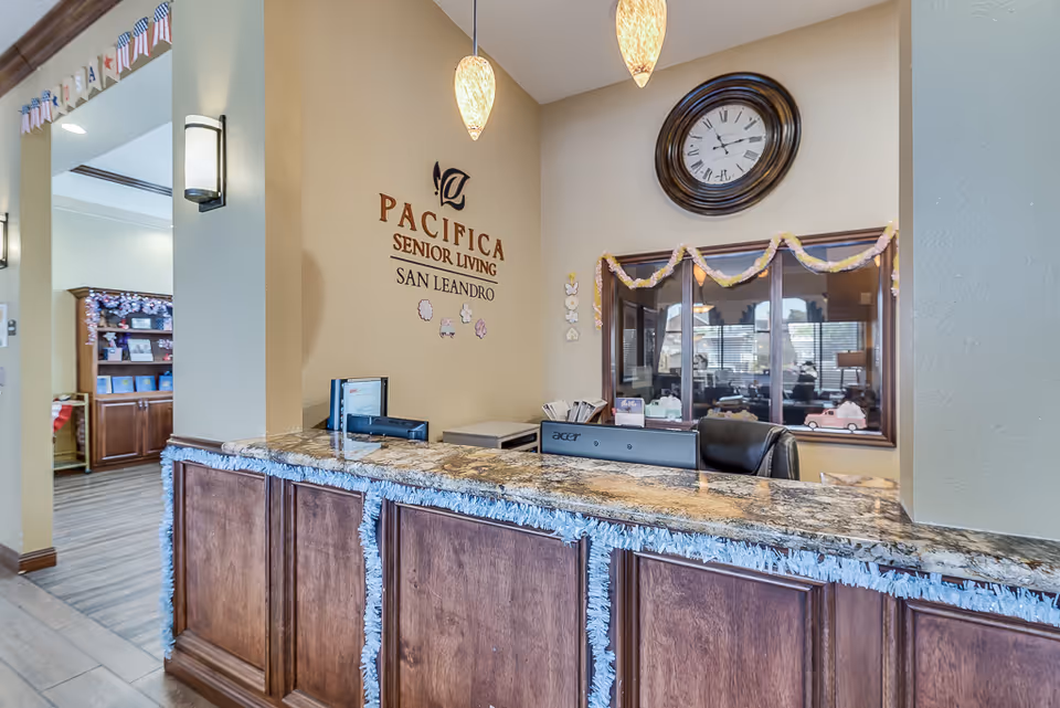 Reception desk area at Pacifica Senior Living San Leandro with a granite countertop decorated with garland. Behind the desk is a wall clock and a window looking into another room. The wall features the facility's name and logo. Pendant lights hang from the ceiling above the desk.