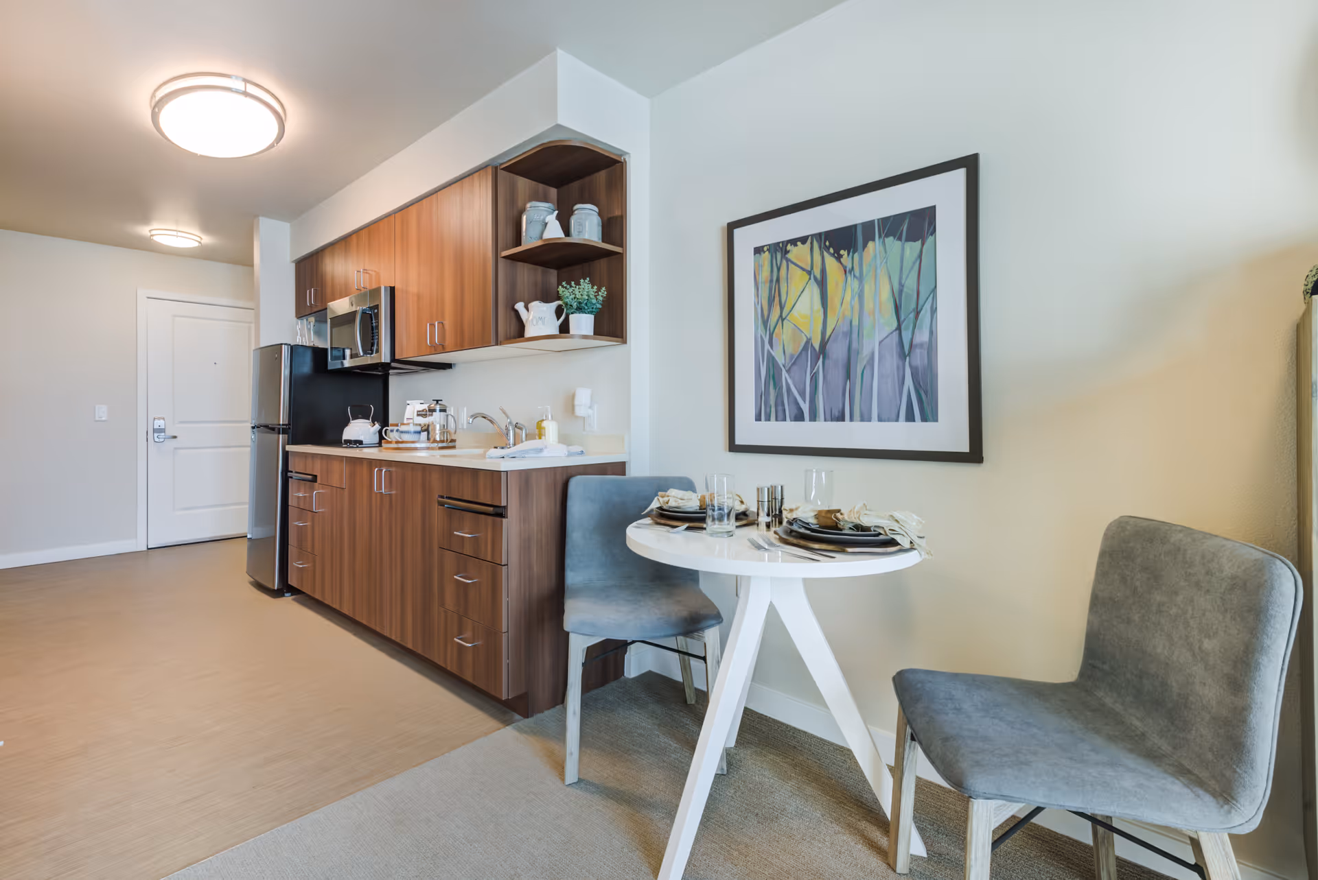 Compact apartment kitchenette with wood cabinets, stainless refrigerator and microwave, and a small round dining table set for two beneath framed artwork.