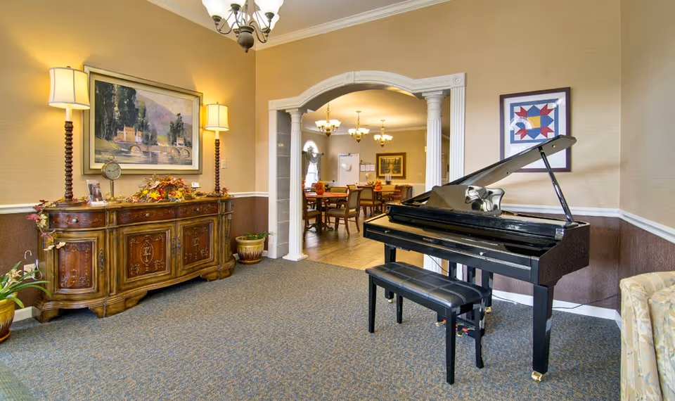 Common room featuring a black grand piano, an ornate wooden sideboard with lamps, and an arched entryway into a dining area.