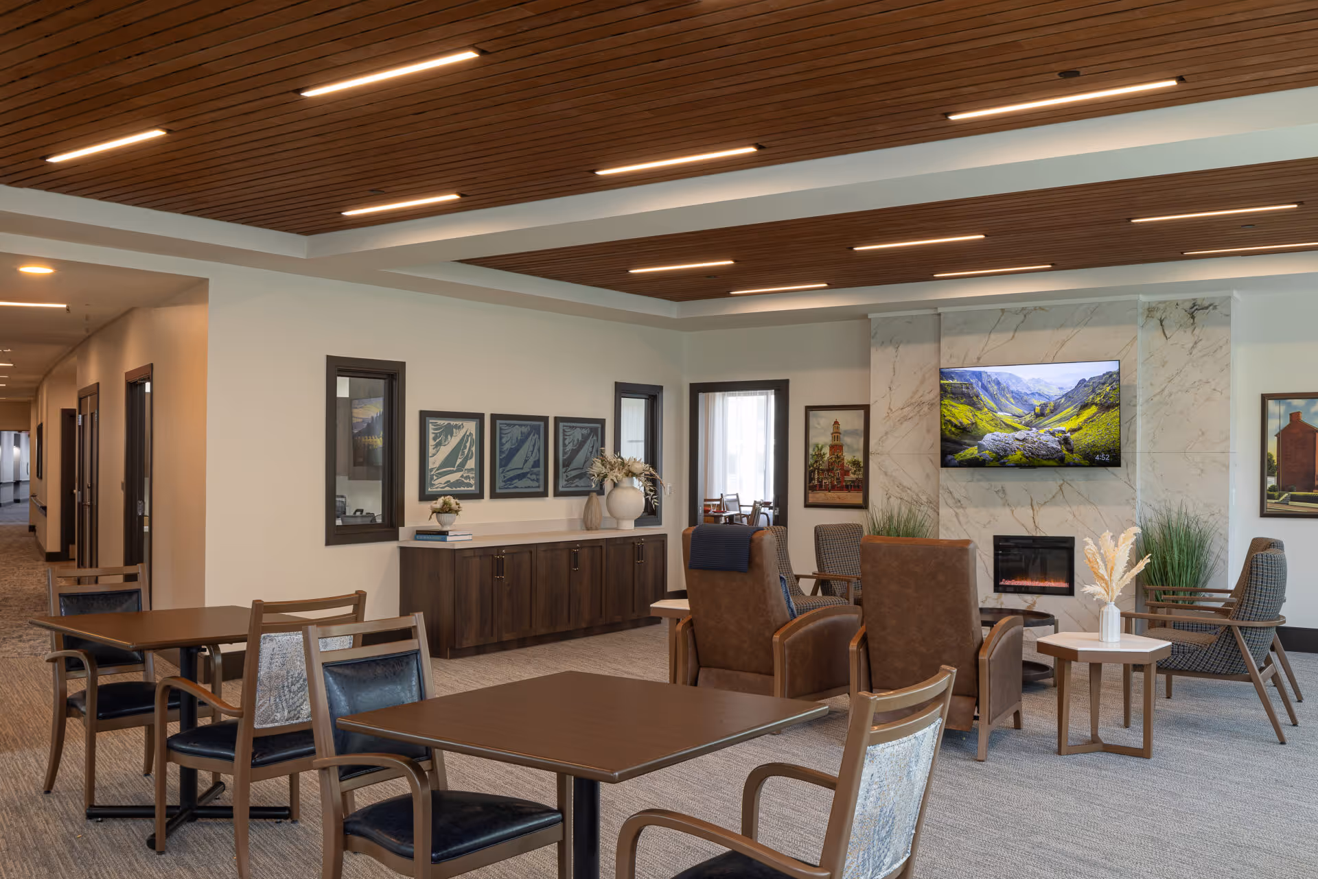 A spacious and modern common area with wooden tables and chairs, comfortable armchairs facing a wall-mounted TV above a fireplace, decorative plants, framed artwork on the walls, and a wooden ceiling with recessed lighting.