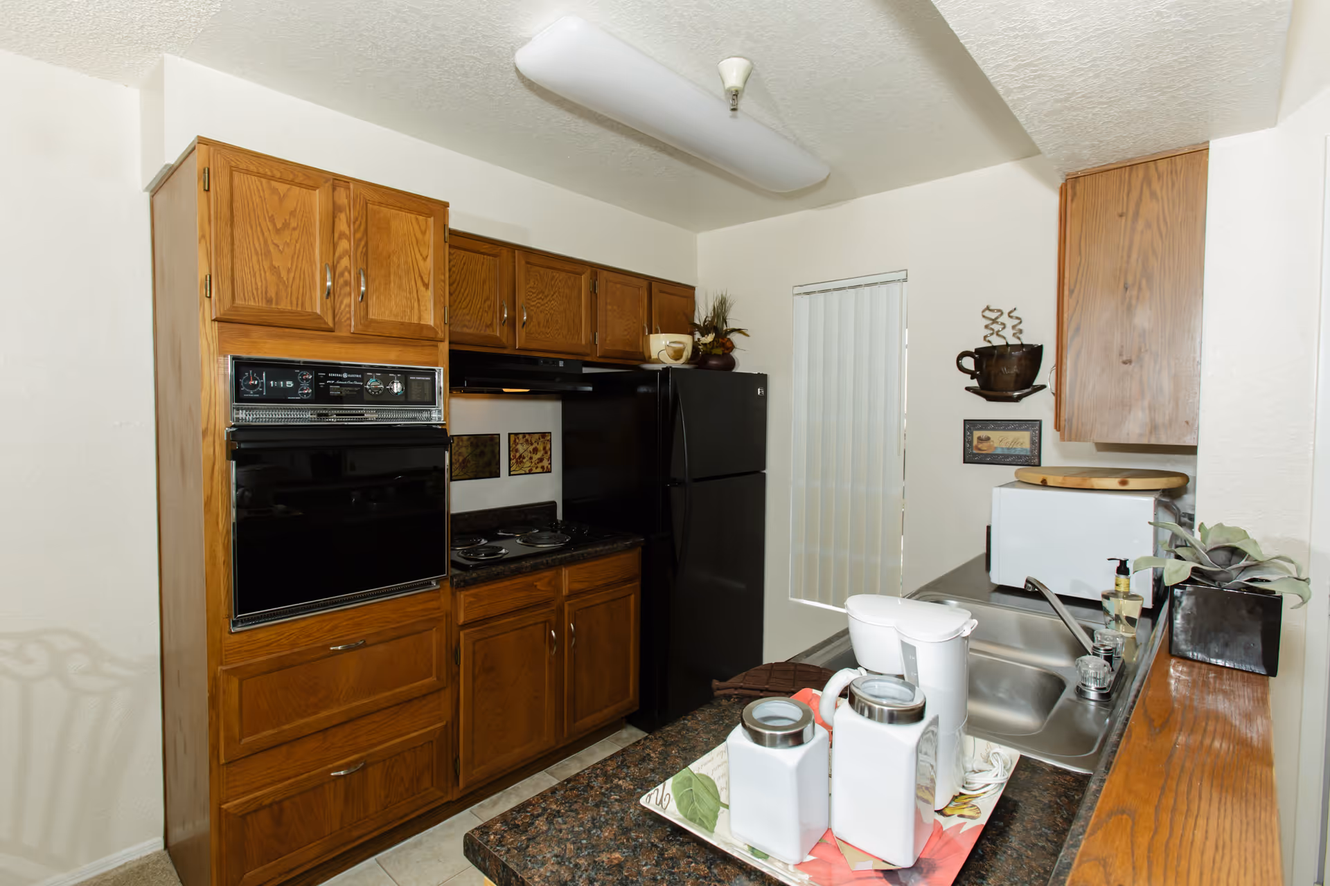 A compact kitchen with wooden cabinets, a black refrigerator, an electric stove with oven, a microwave, and a double sink. The countertop has a tray with two white canisters and a white coffee maker. There is a decorative coffee cup wall hanging and a small plant near the sink.