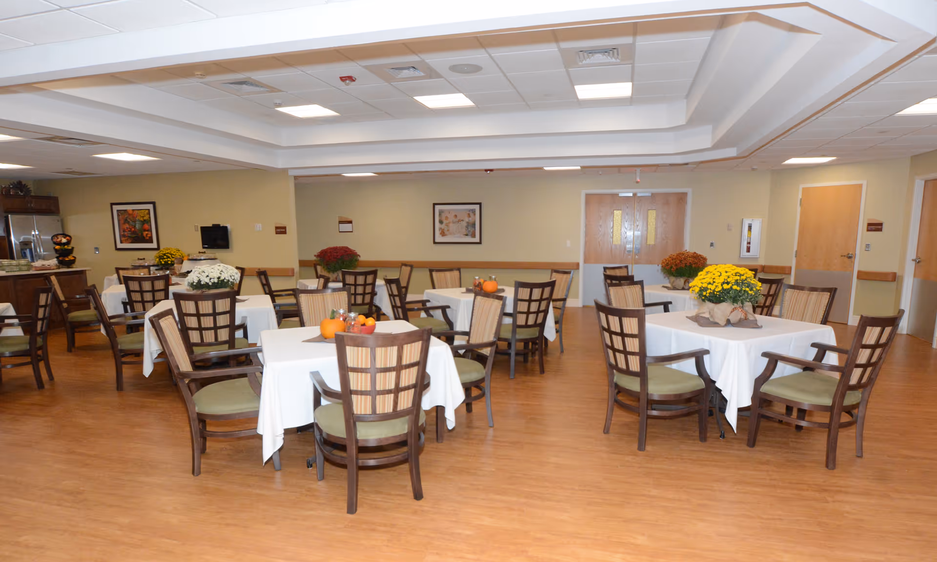 A spacious dining room with several square tables covered with white tablecloths, each surrounded by wooden chairs with green cushions. The tables are decorated with small pumpkins and potted flowers. The room has wood flooring, beige walls, and a white ceiling with recessed lighting. There are double doors and framed artwork on the walls.