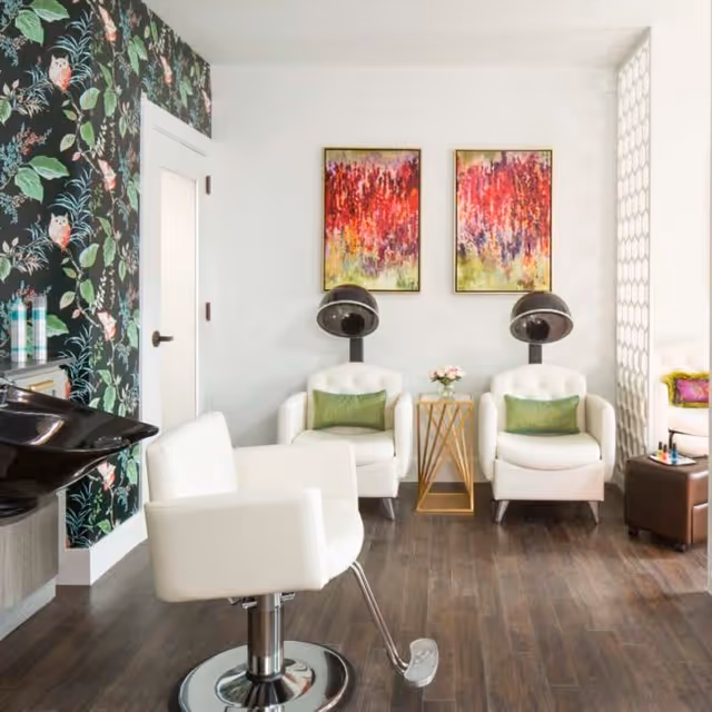 A bright and modern salon area with two white salon chairs facing hair drying stations. The room features dark wood flooring, a floral wallpaper with owls on one wall, two colorful abstract paintings on a white wall, and a small gold side table with a flower vase between the chairs.