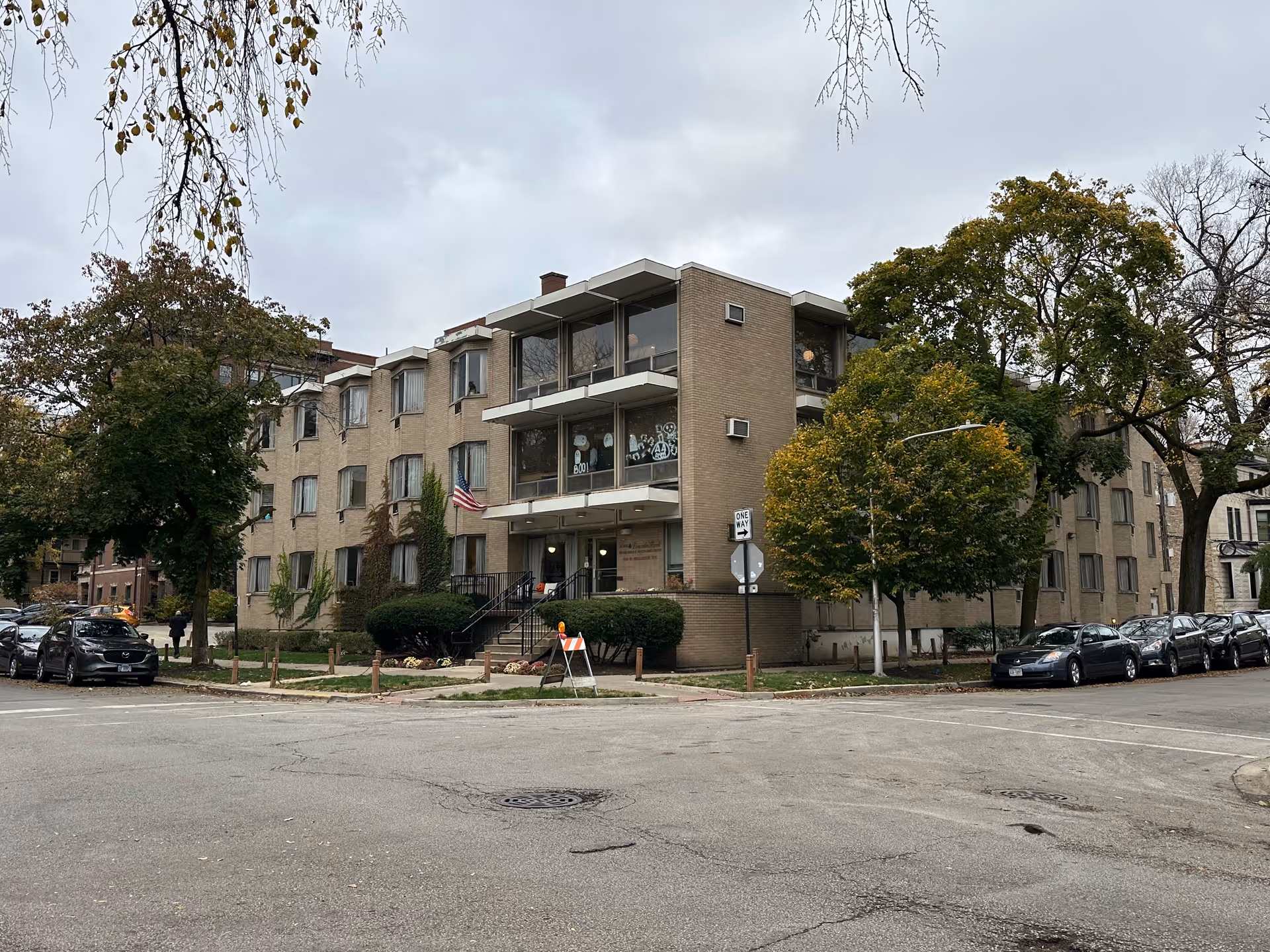 Exterior view of a three-story beige brick building on a street corner with trees and parked cars along the sides. The building has large windows and an American flag near the entrance.