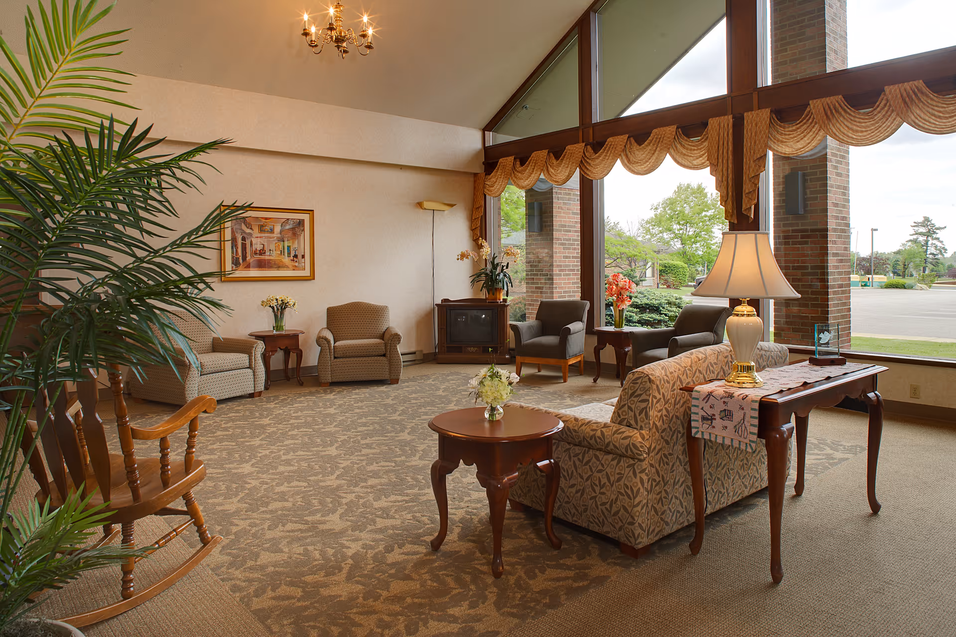 A spacious living room area with large floor-to-ceiling windows letting in natural light. The room features a patterned sofa, several armchairs, wooden side tables with lamps and flower arrangements, a rocking chair, a carpet with a floral design, and a chandelier hanging from the ceiling. Outside the windows, greenery and a parking lot are visible.
