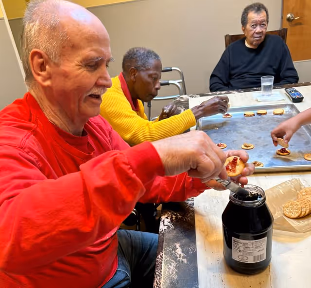 Three elderly individuals sitting around a table preparing crackers with jam. One man in a red shirt is spreading jam on a cracker, a woman in a yellow sweater is placing jam on crackers on a baking tray, and another man in a black shirt is sitting at the table with a glass of water and a remote control nearby.