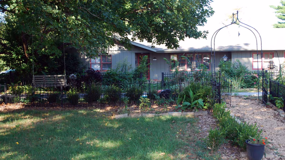 A garden area with a grassy lawn, a black metal fence, and an arched trellis entrance. There are various plants and shrubs along the fence and a wooden bench under a large tree providing shade. In the background, there is a single-story building with gray siding and red window frames.